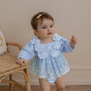 Bebê vestindo vestido azul com flores bordadas, apoiada em cadeira de vime em ambiente neutro.